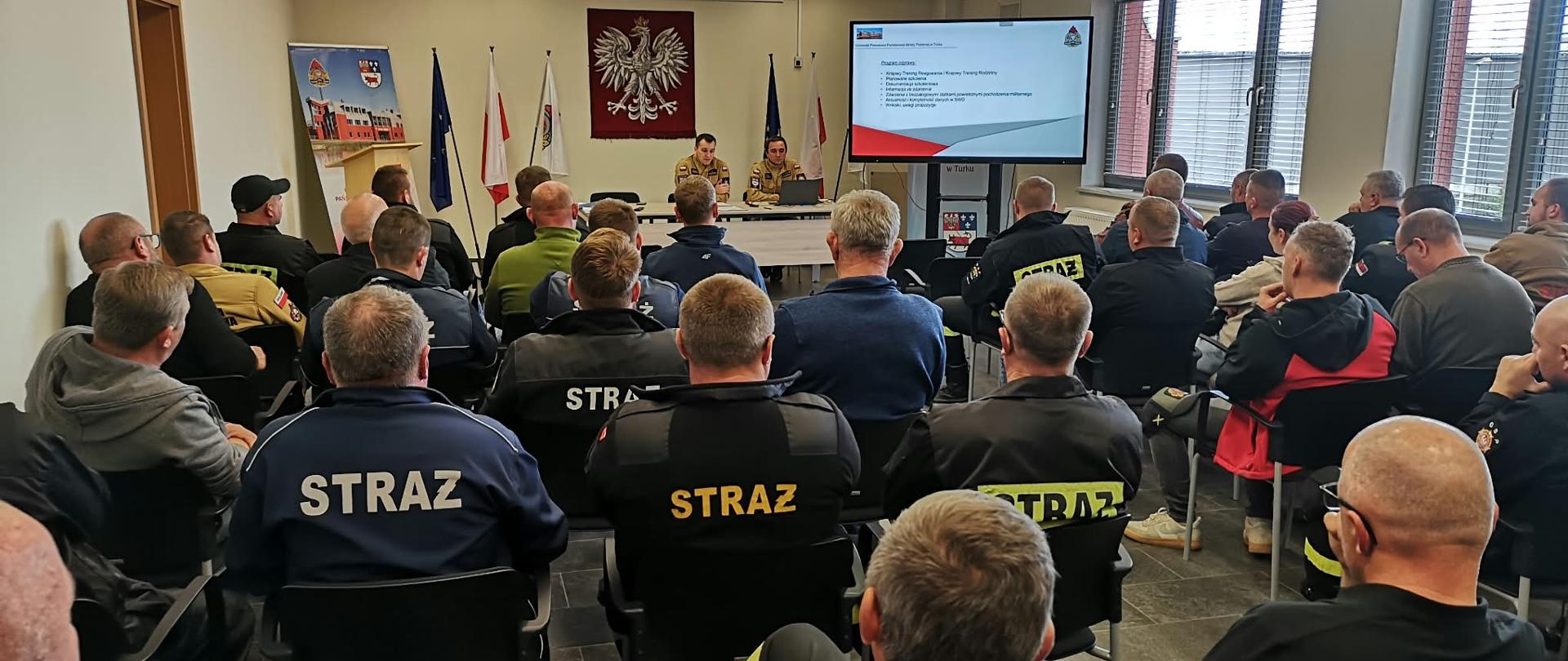 Spotkanie szkoleniowe strażaków w sali konferencyjnej. Uczestnicy siedzą na krzesłach i słuchają prezentacji prowadzonej przez dwie osoby w mundurach