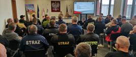 Spotkanie szkoleniowe strażaków w sali konferencyjnej. Uczestnicy siedzą na krzesłach i słuchają prezentacji prowadzonej przez dwie osoby w mundurach