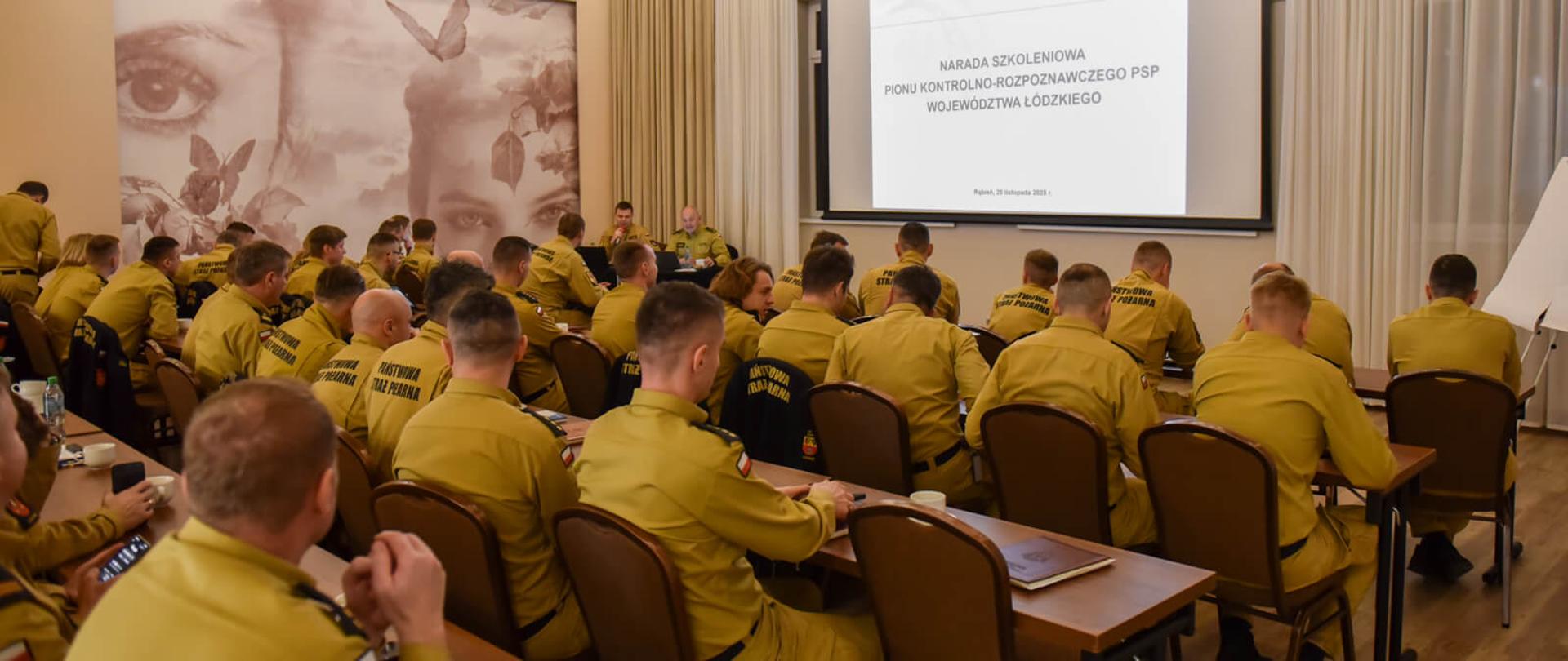Sala konferencyjna z wieloma osobami w mundurach PSP siedzącymi przy stołach i oglądającymi prezentację na dużym ekranie. Na ekranie widać tytuł "NARADA SZKOLENIOWA PIONU KONTROLNO-ROZPOZNAWCZEGO PSP WOJEWÓDZTWA ŁÓDZKIEGO".
