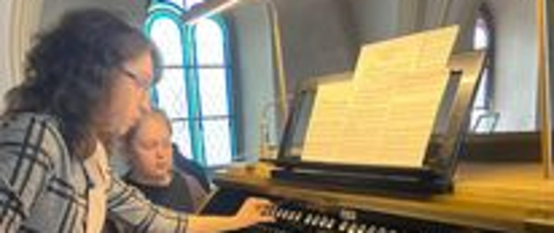 Zdjęcie przedstawiające organy po prawej stronie obrazu, po lewej stronie dwie osoby patrzące na organy.