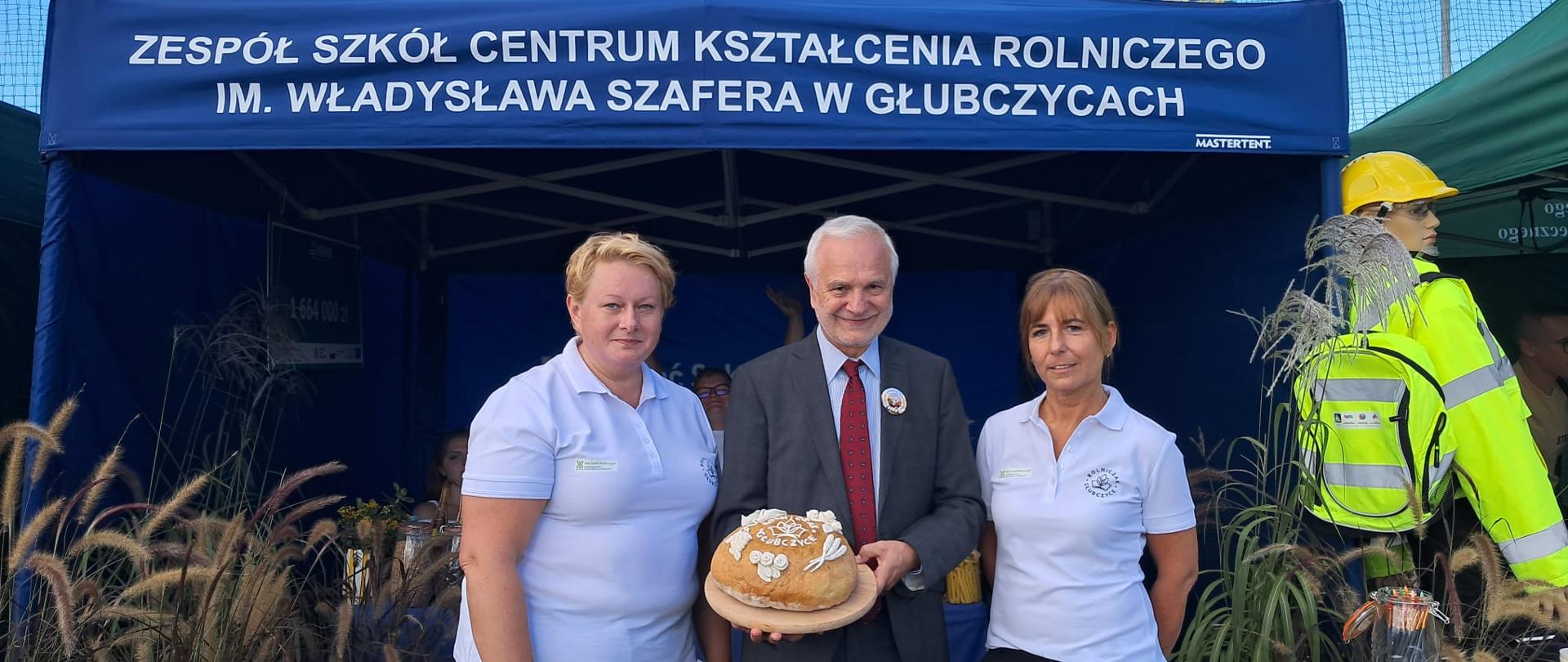 Dyrektor trzyma na desce okrągły chleb ozdobiony napisem „Rolniczak Głubczyce”. Obok stoją dwie nauczycielki w białych koszulkach z logo szkoły, w tle niebieski namiot.