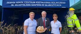 Dyrektor trzyma na desce okrągły chleb ozdobiony napisem „Rolniczak Głubczyce”. Obok stoją dwie nauczycielki w białych koszulkach z logo szkoły, w tle niebieski namiot.