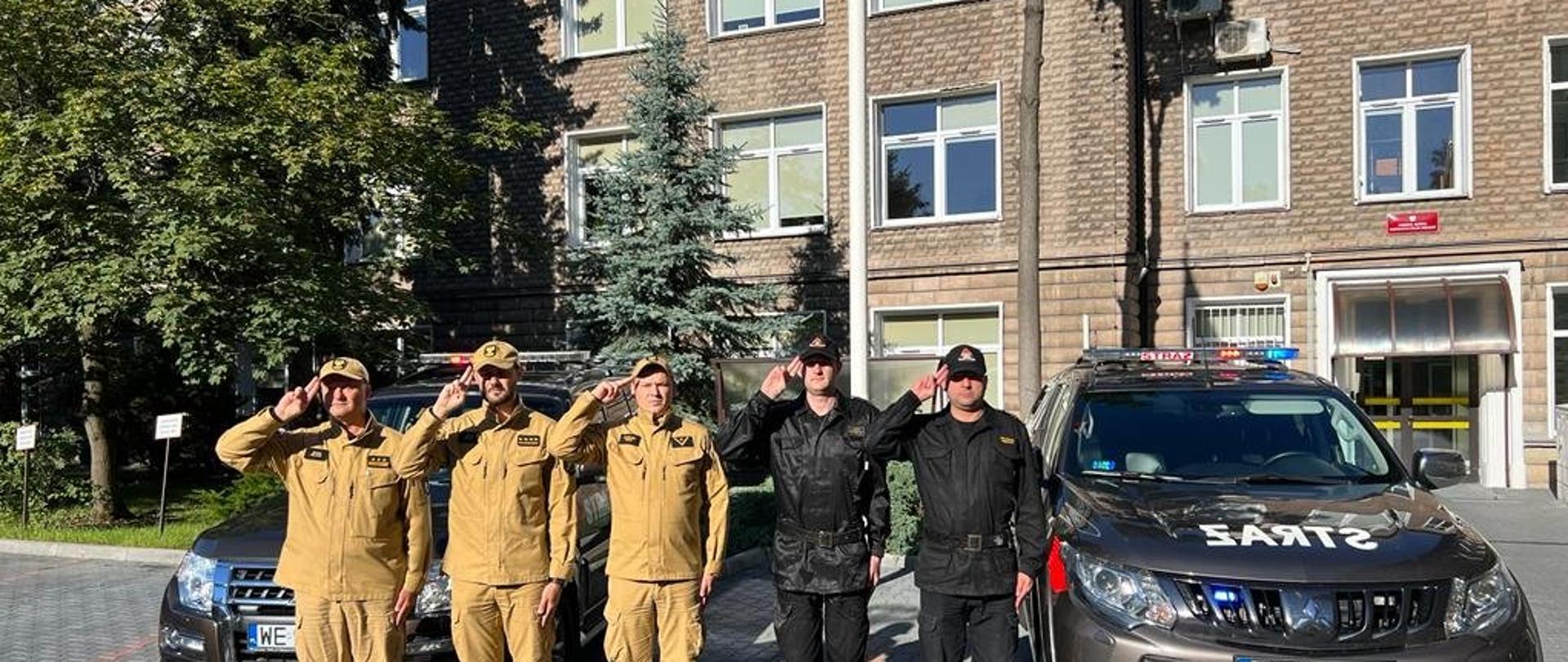 Strażacy przed budynkiem strażnicy stoją na baczność podczas minuty ciszy, za nimi samochody z włączoną sygnalizacją świetlno - dźwiękową