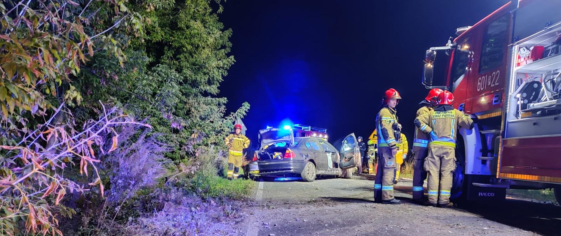Na zdjęciu przy samochodzie strażakom ratownicy ubraniu specjalnym w oddali samochód osobowy na drodze a przy nim również strażacy w ubraniach specjalnych
