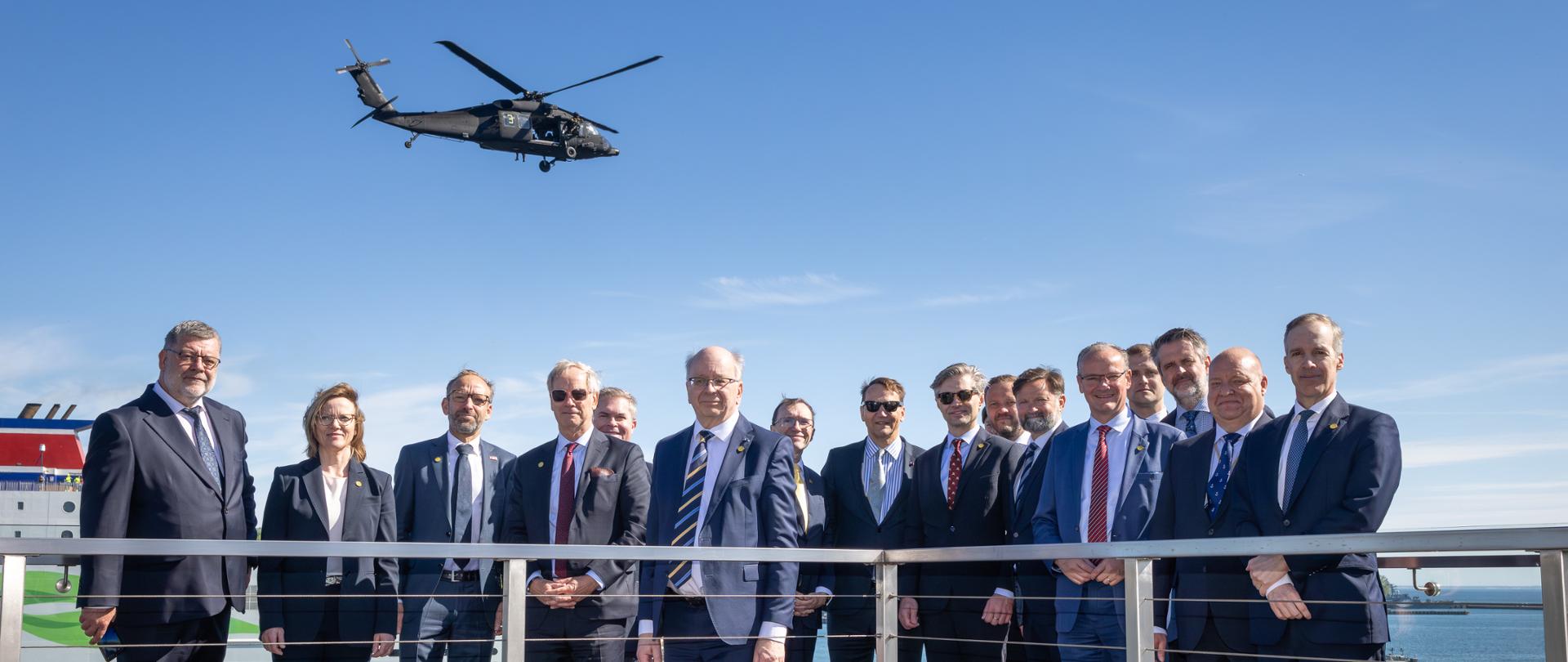 The photo shows a group of people in suits. Behind them is a body of water. Above them is a helicopter. 