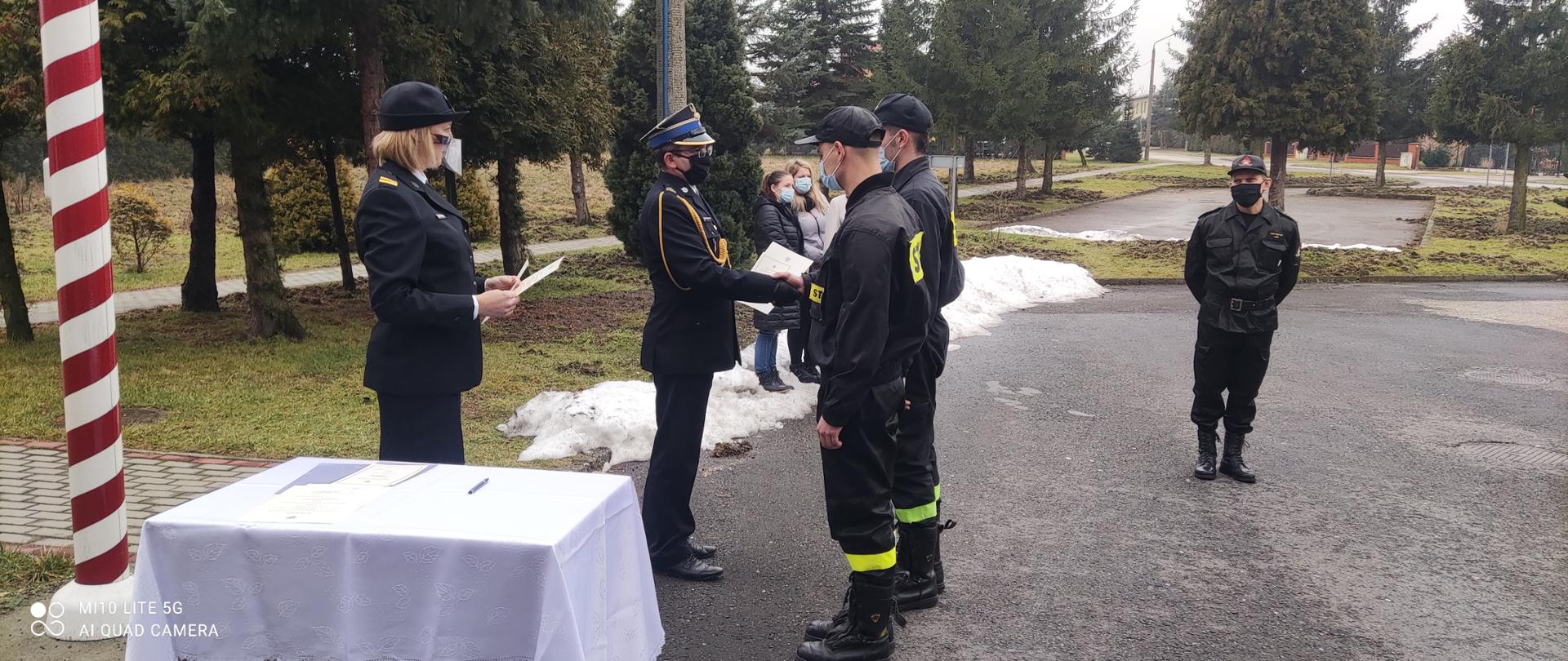 Komendant w mundurze wyjściowym wręcza nowo przyjętym strażakom akty ślubowania. Na pierwszym planie nakryty białym suknem stolik, w tle zaproszeni goście oraz drzewa iglaste.