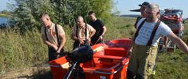 Ćwiczenia z udziałem SGRWN. Zdjęcie przedstawia teren nad zbiornikiem retencyjnym w Jutrosinie. sześciu strażaków przenosi czerwoną łódź ratowniczą, do której zamontowany jest silnik zaburtowy. W tle widoczne są krzewy i drzewa oraz przyczepka ze sprzętem do ratownictwa wodnego.