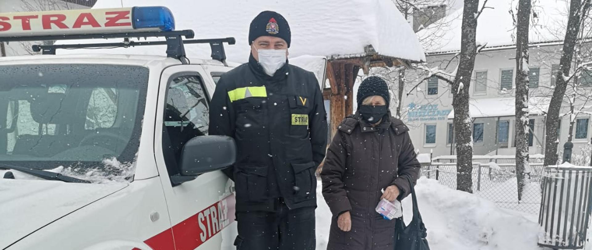 Na zdjęciu widać strażaka w ubraniu koszarowym przy samochodzie terenowym Państwowej Straży Pożarnej. Obok strażaka stoi kobieta z torebką w ręce. W oddali drzewa oraz budynek. Aura jest zimowa.