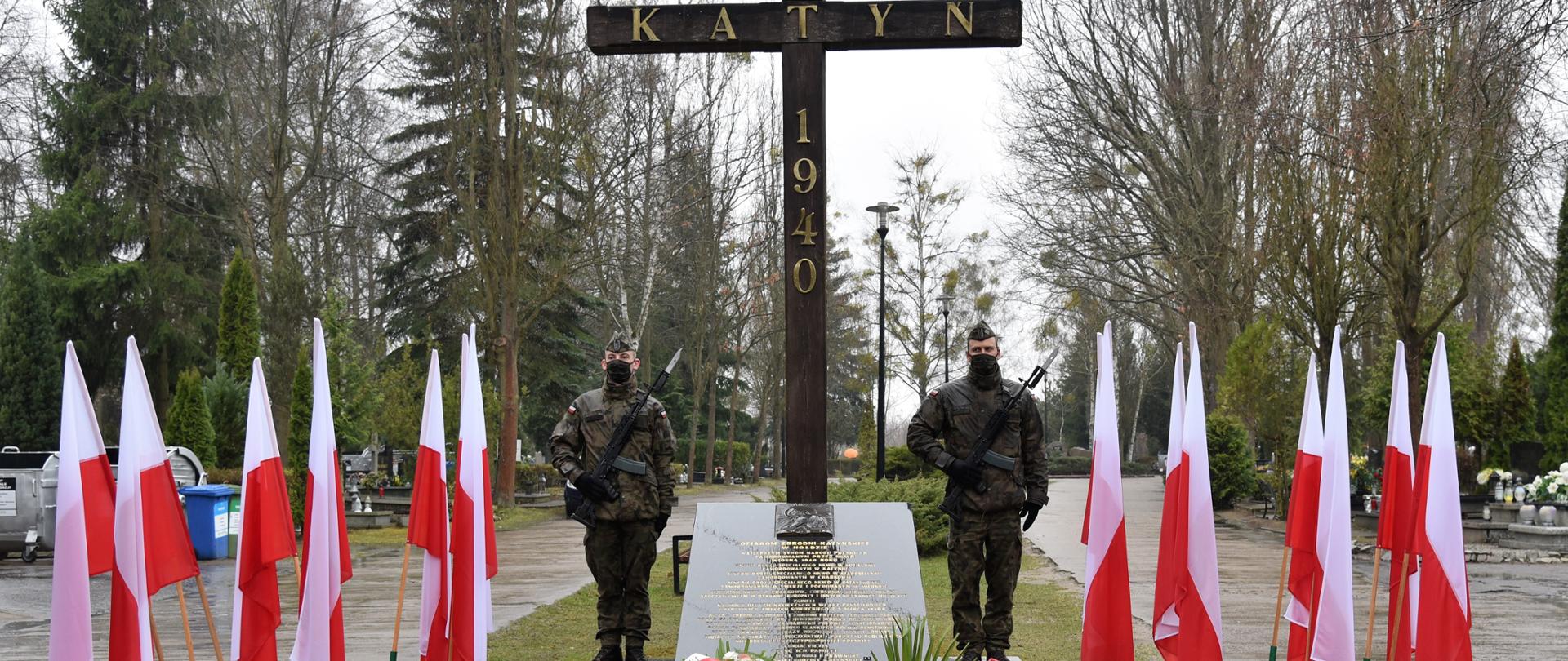 Krzyż ofiar Katynia na którym przybito złocone liczby układające się w datę 1940, pod krzyżem złożone kwiaty i znicze. Obok Krzyża żołnierze trzymają wartę honorową. Po bokach stoją stojaki z biało-czerownymi flagami. W tle główna aleja cmentarza. 