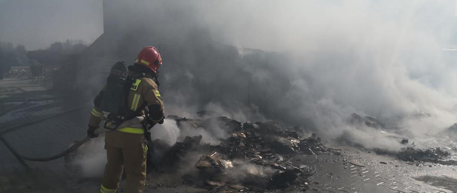 Strażak ubrany w ubranie specjalne i aparat ochrony układu oddechowego podaje prąd wody na tlące się opakowania kartonowe
