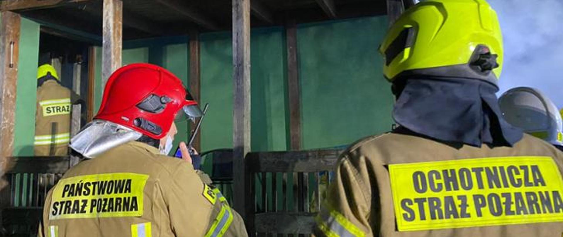 Zdjęcie przedstawia od lewej strażaka Państwowej Straży Pożarnej ubranego w ubranie specjalne oraz czerwony hełm, z prawej strażak Ochotniczej Straży Pożarnej ubrany w ubranie specjalne oraz żółty hełm. W tle budynek w którym prowadzona jest akcja gaśnicza.