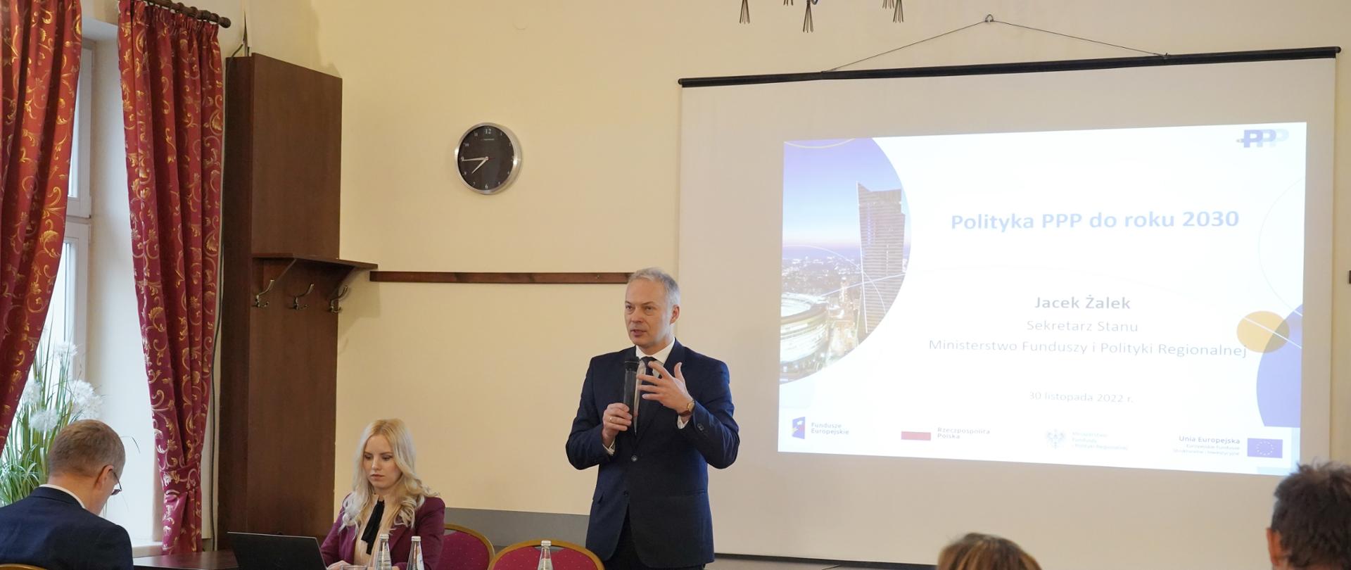  Wiceminister Jacek Żalek stoi przy stoliku i mówi do mikrofonu, który trzyma w prawek dłoni.