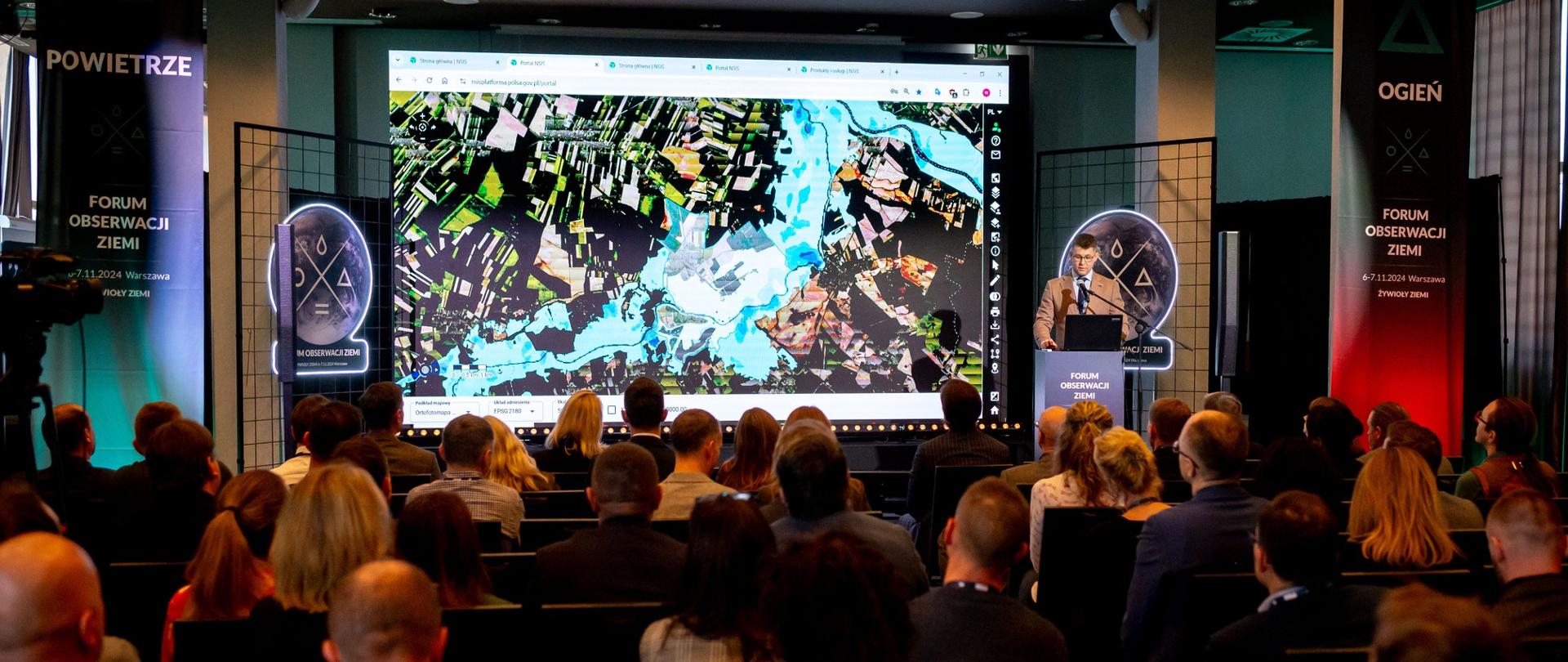 Sala konferencyjna w ciemnych odcieniach. Na pierwszym planie widać ludzi siedzących na krzesłach. W oddali na scenie stoi mężczyzna za mównicą, który przemawia do uczestników Konferencji. Po lewej stronie za mężczyzną na ekranie projekcyjnym wyświetlana jest prezentacja. 