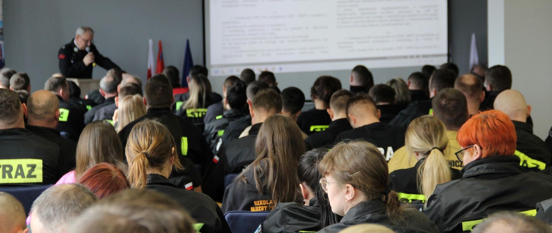 Pełna sala ludzi w mundurach z napisem STRAŻ na plecach. W oddali widać druha z mikrofonem, w tle ekran, a na nim tekst.