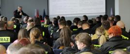 Pełna sala ludzi w mundurach z napisem STRAŻ na plecach. W oddali widać druha z mikrofonem, w tle ekran, a na nim tekst.