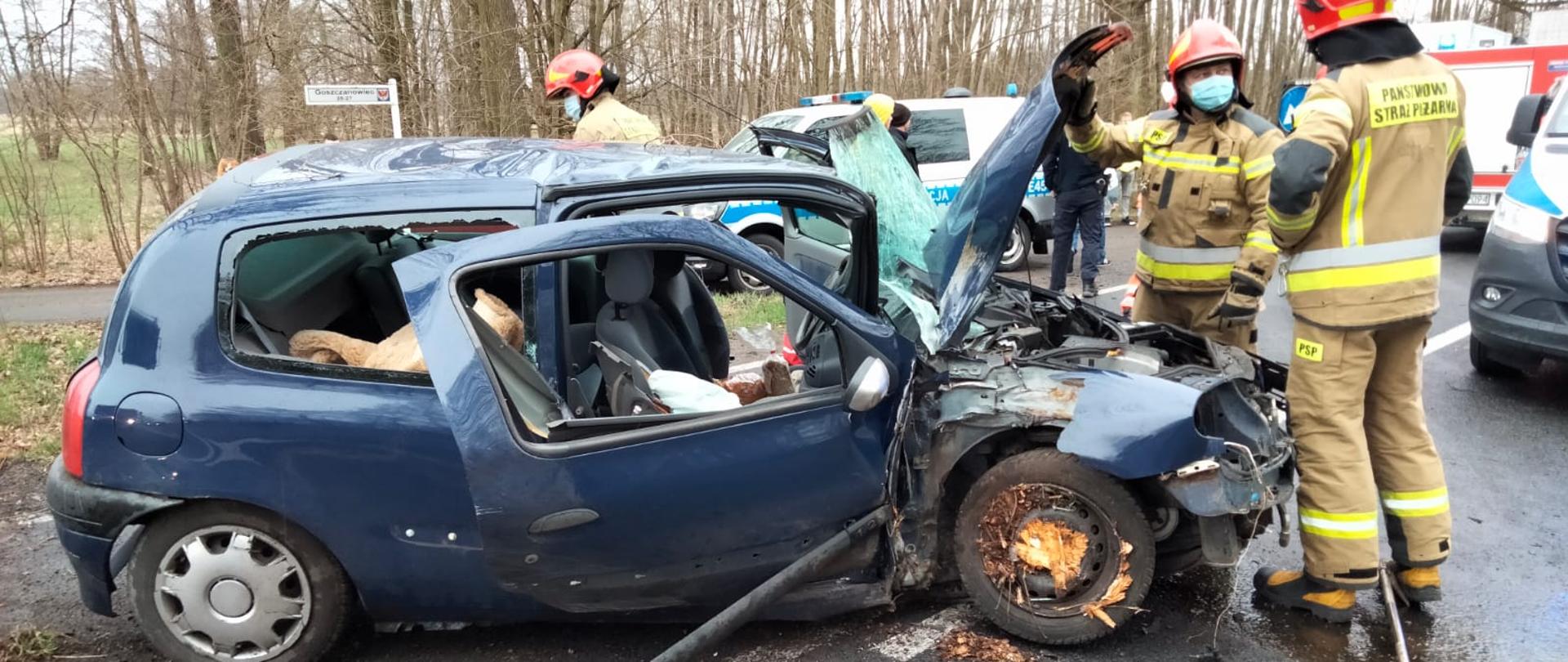 Fotografia przedstawia ujęcie prawego boku samochodu osobowego marki renault clio koloru granatowego. Szyby w pojeździe są wybite, zniszczone jest prawe przednie nadkole, na stalowej feldze przedniego prawego koła widoczne duże ilości pozdzieranej kory drzewa. Pojazd stoi na asfalcie, przy otwartej masce stoi dwóch strażaków w ubraniach specjalnych i w hełmach, jeden stoi tyłem, drugi przodem do fotografującego; strażak stojący przodem na twarzy ma maseczkę chirurgiczną. Za pojazdem widoczny trzeci strażak, w tle widoczne pojazdy straży pożarnej, policji oraz zespołu ratownictwa medycznego, dalej w tle widać drzewa. Jest dzień.