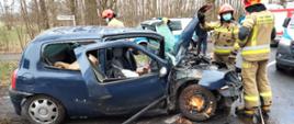 Fotografia przedstawia ujęcie prawego boku samochodu osobowego marki renault clio koloru granatowego. Szyby w pojeździe są wybite, zniszczone jest prawe przednie nadkole, na stalowej feldze przedniego prawego koła widoczne duże ilości pozdzieranej kory drzewa. Pojazd stoi na asfalcie, przy otwartej masce stoi dwóch strażaków w ubraniach specjalnych i w hełmach, jeden stoi tyłem, drugi przodem do fotografującego; strażak stojący przodem na twarzy ma maseczkę chirurgiczną. Za pojazdem widoczny trzeci strażak, w tle widoczne pojazdy straży pożarnej, policji oraz zespołu ratownictwa medycznego, dalej w tle widać drzewa. Jest dzień.