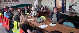 Zdjęcie przedstawia grupę ludzi siedzących przy stołach konferencyjnych w sali wykładowej lub szkoleniowej. Na ścianie w tle widoczne jest duże, charakterystyczne logo firmy KGHM Polska Miedź. Widać przewieszone przez krzesła jaskrawe kamizelki odblaskowe, a na stołach znajdują się dokumenty oraz napoje.