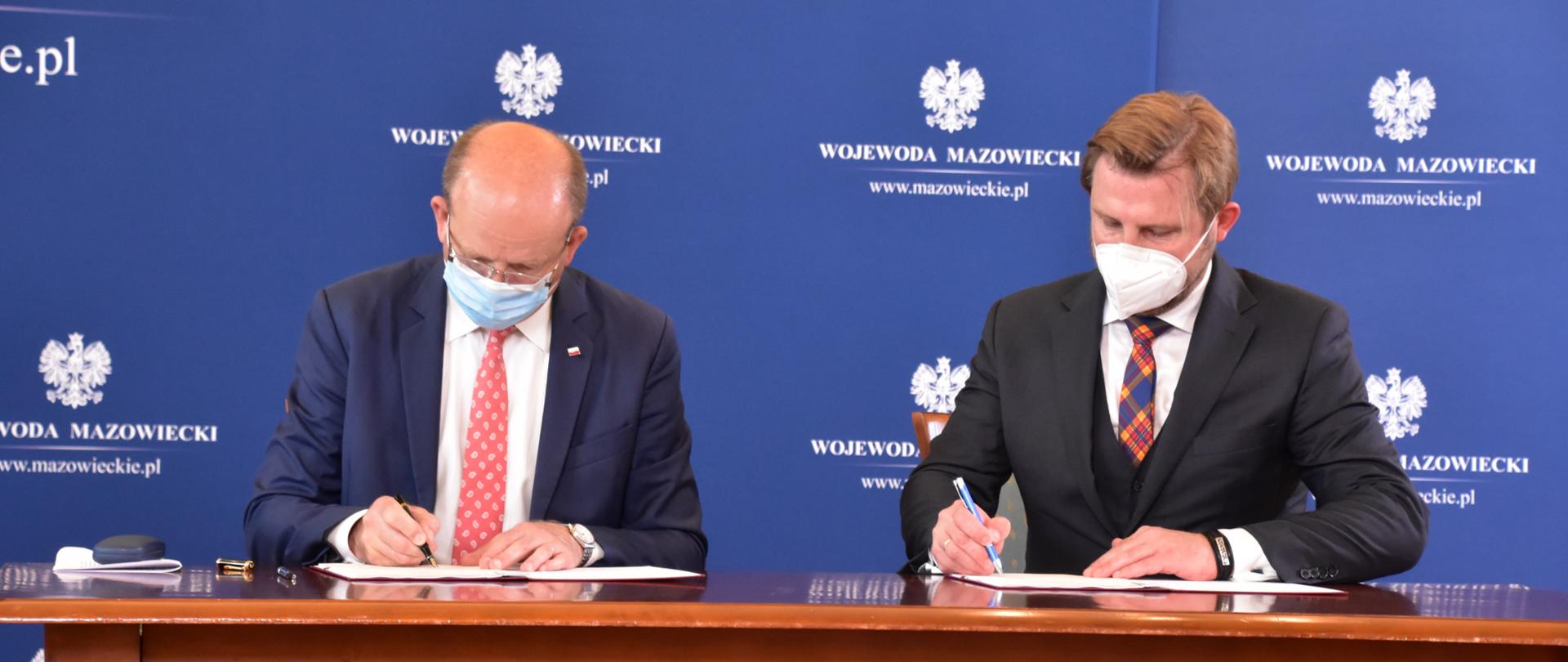 Zdjęcie przedstawia moment podpisania umowy na dofinansowanie utworzenia Centrum opiekuńczo-mieszkalnego w Starych Babicach. Na zdjęciu znajduje się dwóch, ubranych w garnitury mężczyzn. Mężczyźni Ci siedzą za stołem. W tle znajduje się granatowa ścianka wizerunkowej Wojewody Mazowieckiego.