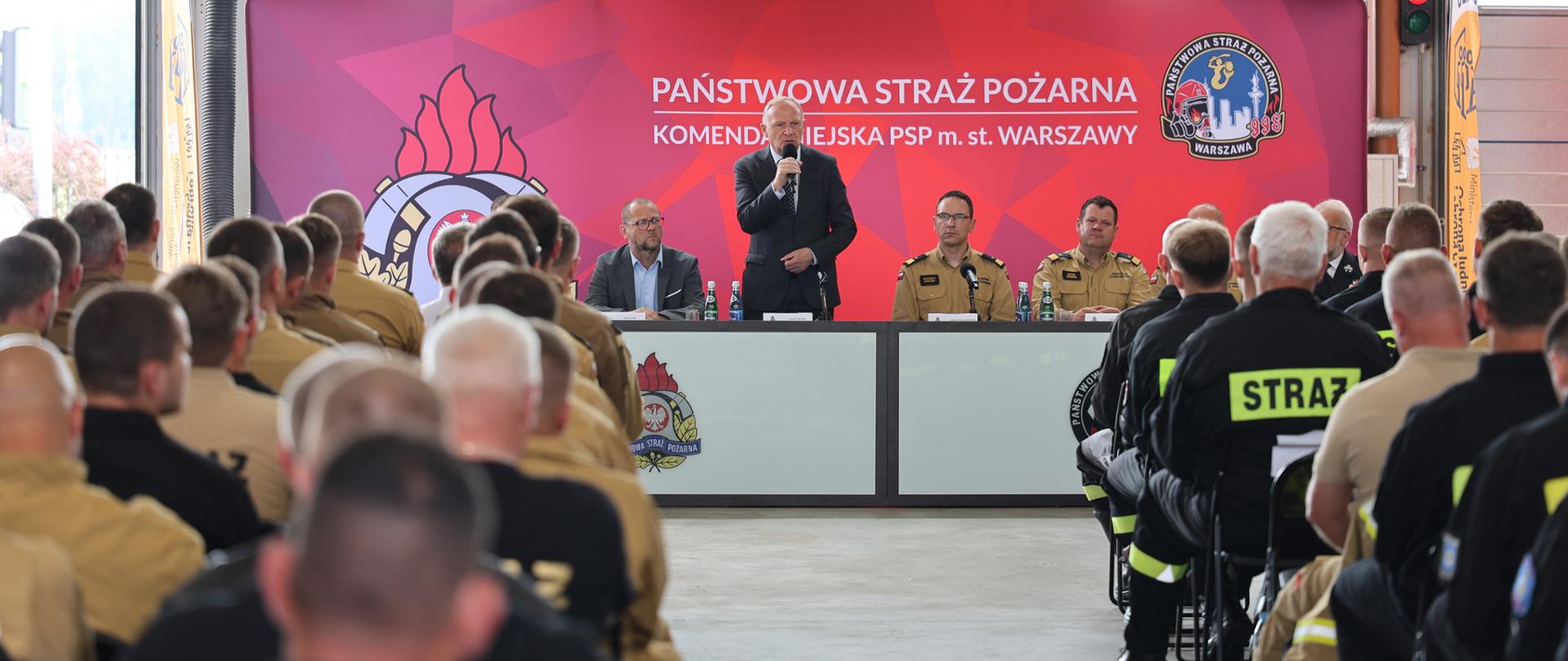 Zdjęcie przedstawia salę w remizie lub hali garażowej podczas konferencji. Na pierwszym planie widoczne są plecy uczestników siedzących na krzesłach – część z nich ma piaskowe mundury PSP, a część czarne ubrania z odblaskowymi napisami „STRAŻ”. W głębi znajduje się scena z prelegentami siedzącymi za stołem na tle czerwonej ścianki z logo Państwowej Straży Pożarnej. Nad sceną widoczny jest ekran prezentacyjny.