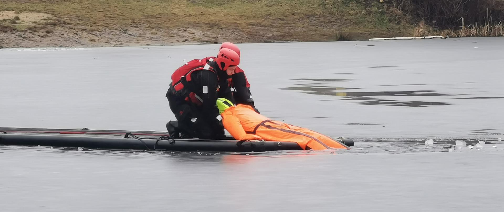 Na zamarzniętym zbiorniku wodnym dwóch strażaków na pneumatycznym trapie/podeście ratowniczym wyciąga z wody manekin, który imituje ofiarę wypadku