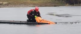 Na zamarzniętym zbiorniku wodnym dwóch strażaków na pneumatycznym trapie/podeście ratowniczym wyciąga z wody manekin, który imituje ofiarę wypadku
