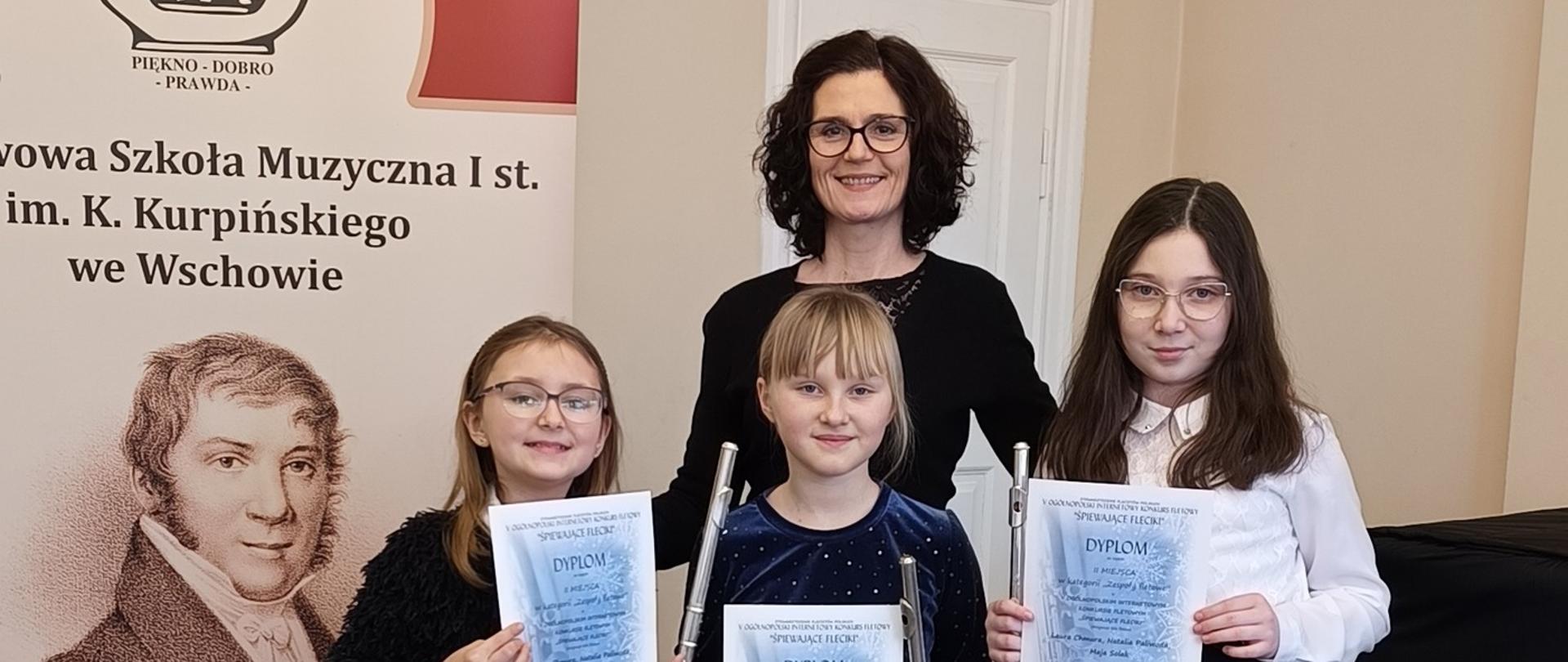 Nagrodzone uczennice wraz z nauczycielką. Zdjęcie wykonane w auli szkoły. Uczennice trzymają w dłoniach dyplomy oraz instrumenty.
