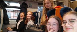 Uśmiechnięte dziewczęta znajdują się w pociągu. Czwórka z nich siedzi, jedna stoi w przejściu 