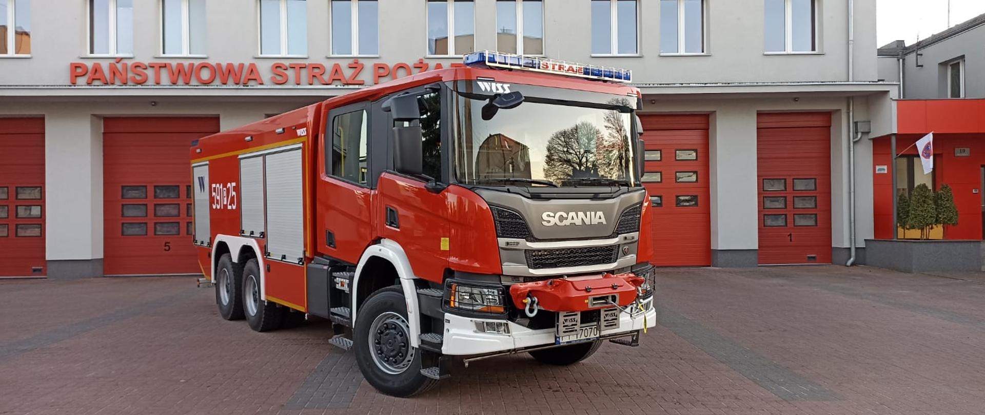 Widać czerwony samochód strażacki Scania od przodu i prawego boku na tle budynku komendy straży pożarnej, za nim czerwone bramy garażowe