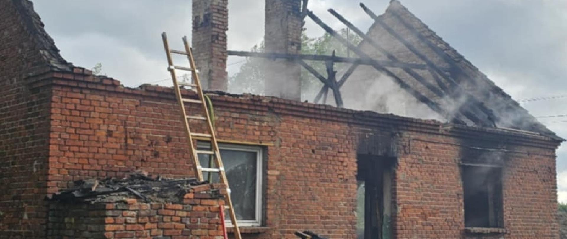 Zdjęcie przedstawia budynek, w którym doszło do pożaru. Widoczne elementy spalonej konstrukcji dachu i zadymienie wydobywające się otworami okiennymi.