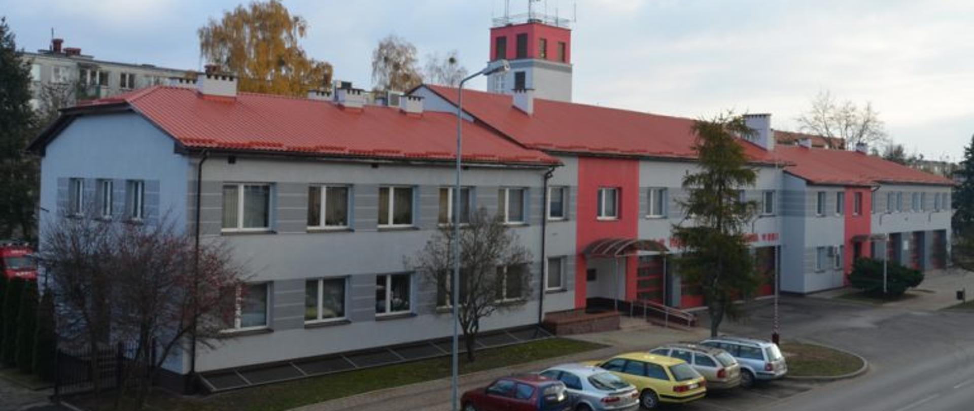 Budynek jednopiętrowy z wysokim parterem rozmieszczonym w prawej części obiektu. Budynek posiada dach dwuspadowy. W części z wysokim parterem widoczne są bramy garażowe. Na parterze i piętrze widoczne okna. Za budynkiem widoczny jest wąski i wysoki budynek, na którym umieszczono anteny radiowe. Przed budynkiem znajduje się ulica oraz chodnik dla pieszych, pomiędzy którymi zaparkowane zostało pięć samochodów osobowych. Przed budynkiem widoczne są również pojedyncze drzewa i krzewy ozdobne. 