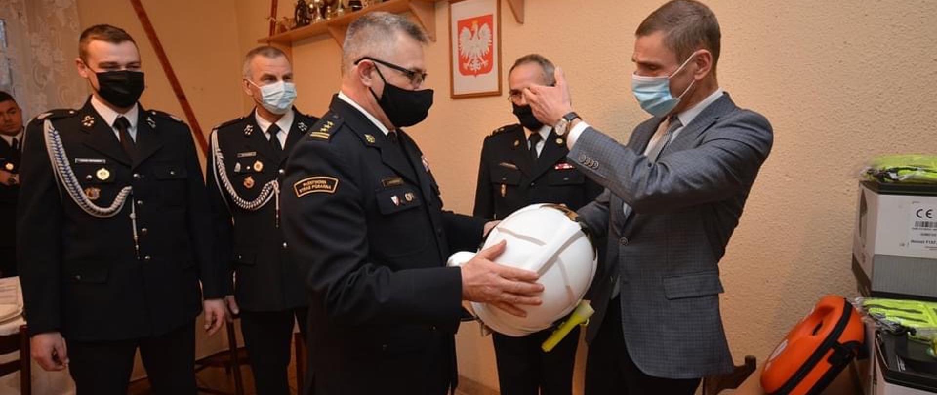 Strażacy wraz z komendantami i burmistrzem. Po prawej stronie przekazywany sprzęt. Burmistrz podaje Kujawsko-Pomorskiemu Komendantowi Państwowej Straży Pożarnej hełm koloru białego.