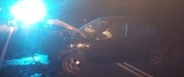 Uszkodzony samochód osobowy stojący bokiem na środku jezdni. Uszkodzony przód samochodu. Zdjęcie wykonane w porze nocnej. W tle blask świateł.