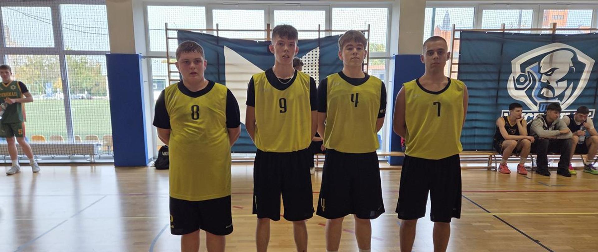 Czterej młodzi koszykarze pozują do zdjęcia stojąc na boisku w hali sportowej.
