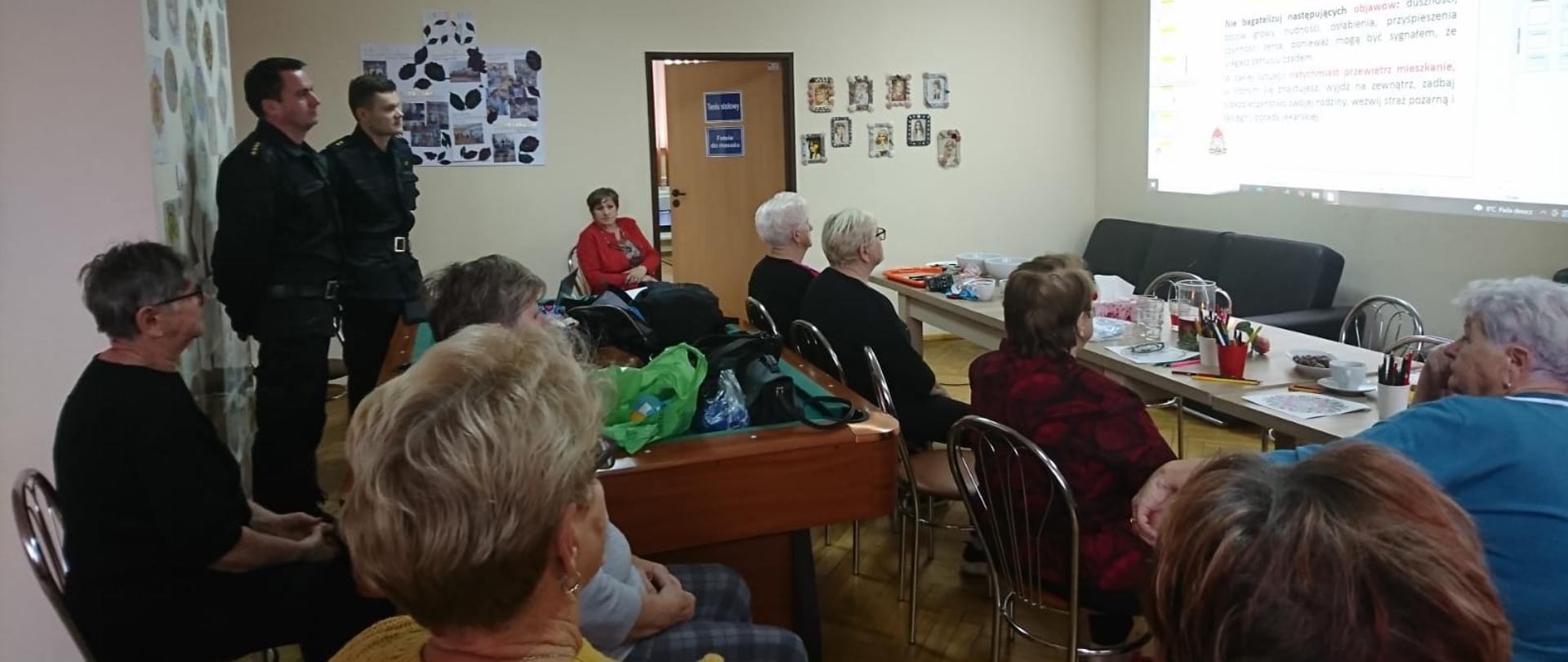 Na zdjęciu na drugim planie dwóch funkcjonariuszy Państwowej Straży Pożarnej prowadzi pogadankę dla seniorów. Pozostali uczestniczy to seniorzy klubu Biedroneczki, którzy zajmują miejsca siedzące i obserwują wyświetlaną na ścianie prezentację.