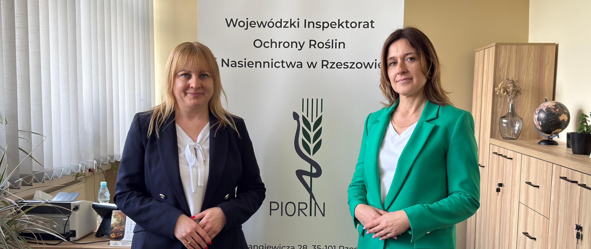 Dwie kobiety stoją obok siebie w biurze na tle roll-upu z napisem instytucji „Wojewódzki Inspektorat Ochrony Roślin i Nasiennictwa w Rzeszowie” oraz logo PIORiN. Kobieta po lewej ma jasne włosy i ubrana jest w granatowy garnitur z białą bluzką, trzyma dłonie złożone przed sobą. Kobieta po prawej ma ciemniejsze włosy i nosi zielony garnitur z białą bluzką, stoi w podobnej, formalnej pozie. W tle widoczne są meble biurowe, rośliny doniczkowe oraz sprzęt biurowy. Atmosfera jest oficjalna i profesjonalna.