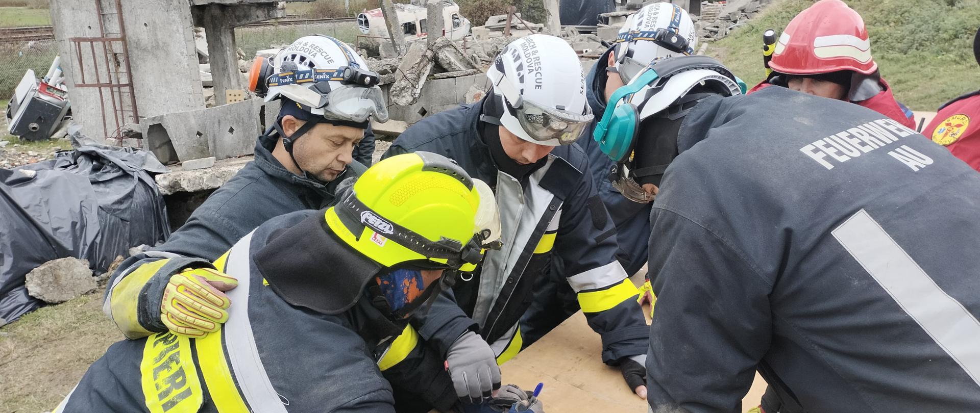 6 strażaków w strojach strażackich i hełmach pochyla się nad stołem, gdzie zapisują coś w notesie. Z tyłu widać zawalony budynek