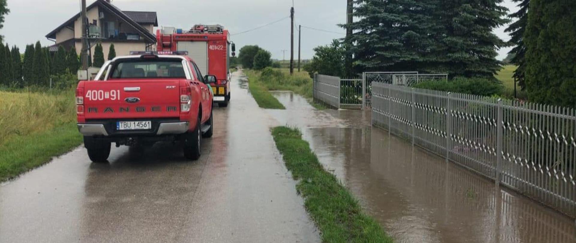 2 samochody pożarnicze jeden za drugim stojące jeden przez drugim na asfalcie. Po lewej stronie woda w rowie. W tle dom