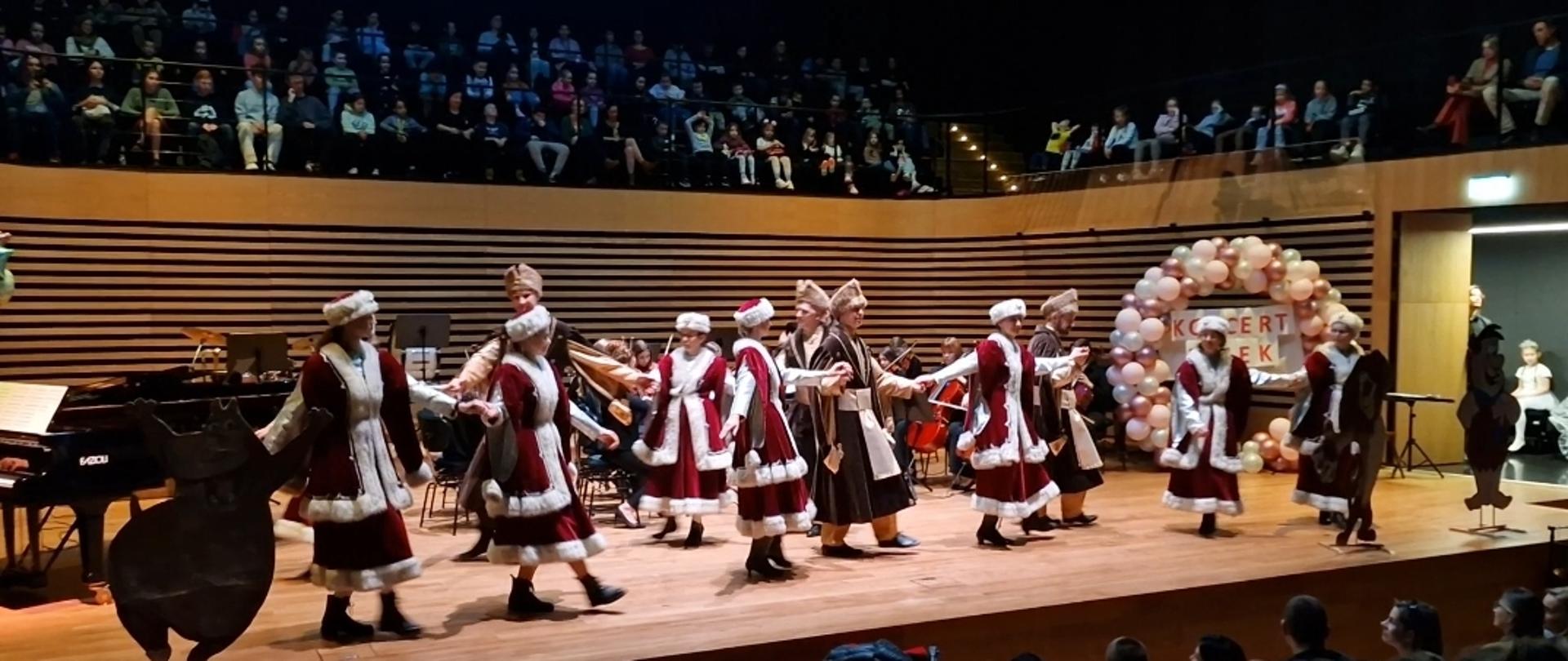Siedem kobiet i czwórka mężczyzn tańczą w szlacheckich strojach na scenie sali koncertowej, z przodu i nad sceną widać publiczność.