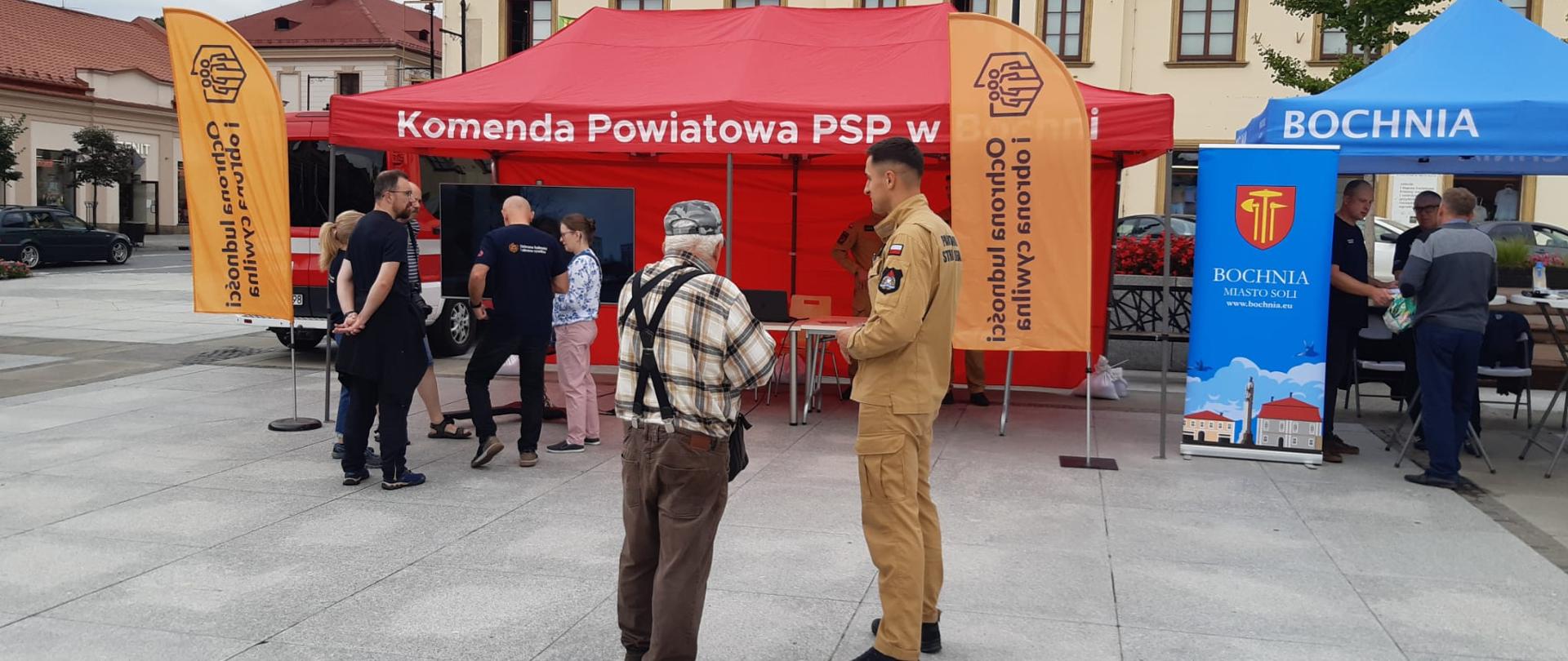 Stoisko Komendy Powiatowej PSP w Bochni podczas wydarzenia plenerowego. Strażacy w mundurach prezentują stanowisko edukacyjne, przy którym rozmawiają z uczestnikami. W tle widoczne są samochody pożarnicze oraz ekran multimedialny.