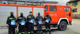 Dystrybucja ulotek. Pięciu strażaków OSP na tle samochodu strażackiego. Strażacy trzymają w ręku ulotki.