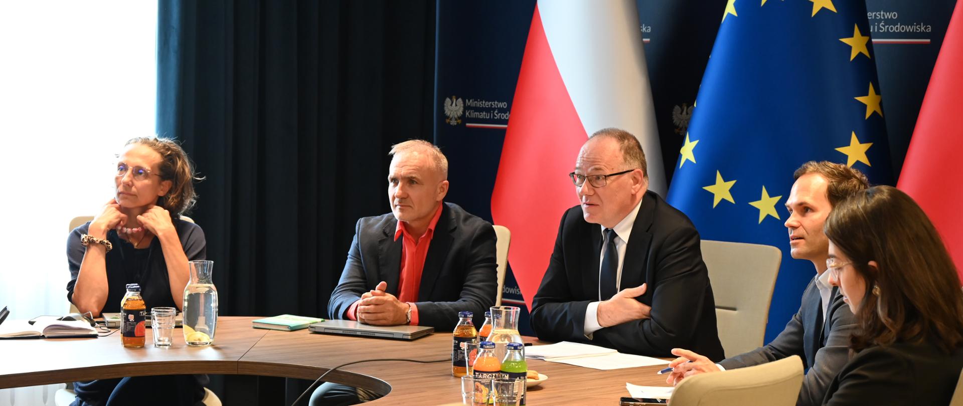 Główny Geolog Kraju Krzysztof Galos podczas spotkania z zastępczynią ministra Isabellą Chan, przedstawicielką kierownictwa Ministerstwa Zasobów Naturalnych Kanady. Na zdjęciu wiceminister siedzi przy okrągłym stole. Za nim widać flagę Polski oraz flagę Unii Europejskiej.