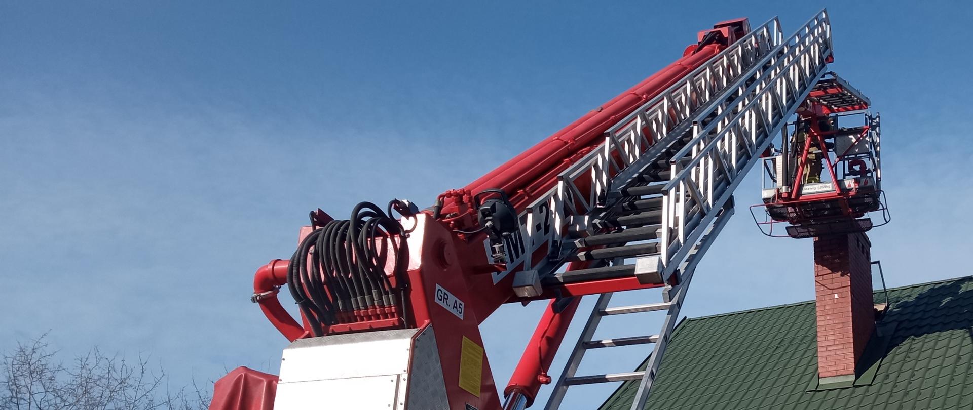 Zdjęcie w ciągu dnia. Widać samochód straży pożarnej. Samochód w kolorze czerwonym wraz z sygnalizacją ostrzegawczą. Lekkie zachmurzenie. W tle widać drzewa. Po prawej fragment budynku jednorodzinnego z zielonym dachem.