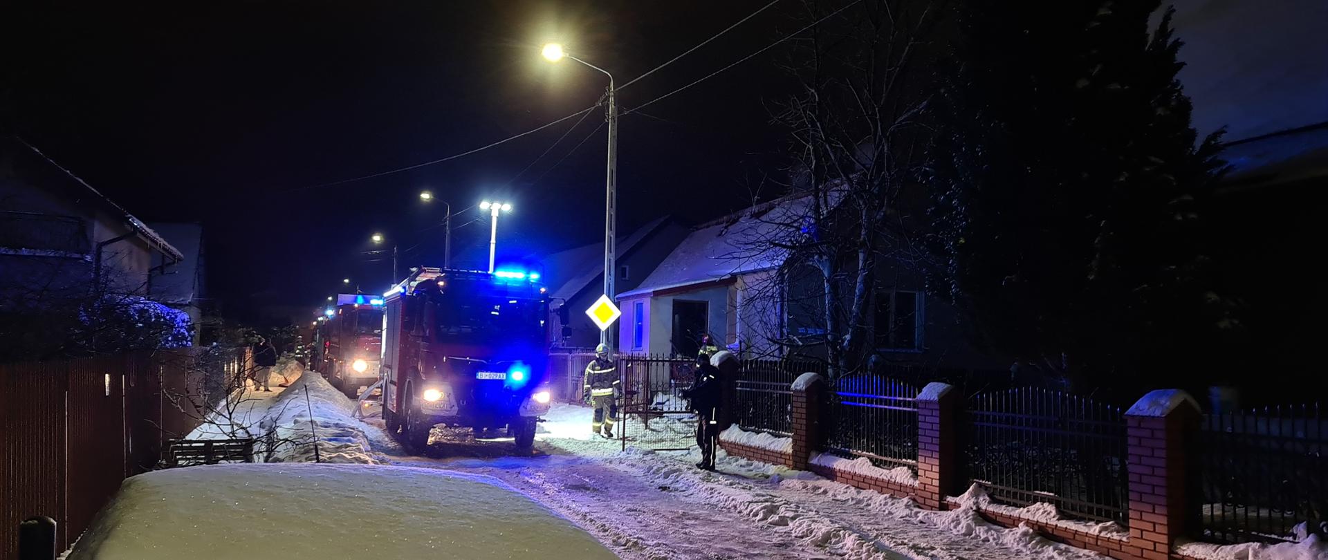 Zdjęcie przedstawia strażaków podczas prowadzenia akcji ratowniczo-gaśniczej na ulicy Słonecznej w Zambrowie. W centralnej części zdjęcia zrobionego w zimową noc stoją samochody jednostek Straży Pożarnej na sygnałach. Na zdjęciu po jego prawej stronie widać dom jednorodzinny oraz strażaków w ubraniach bojowych podczas prowadzenia działań.