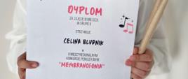 Zdjęcie przedstawia uczennicę trzymającą dyplom oraz pałki perkusyjne na białym tle.