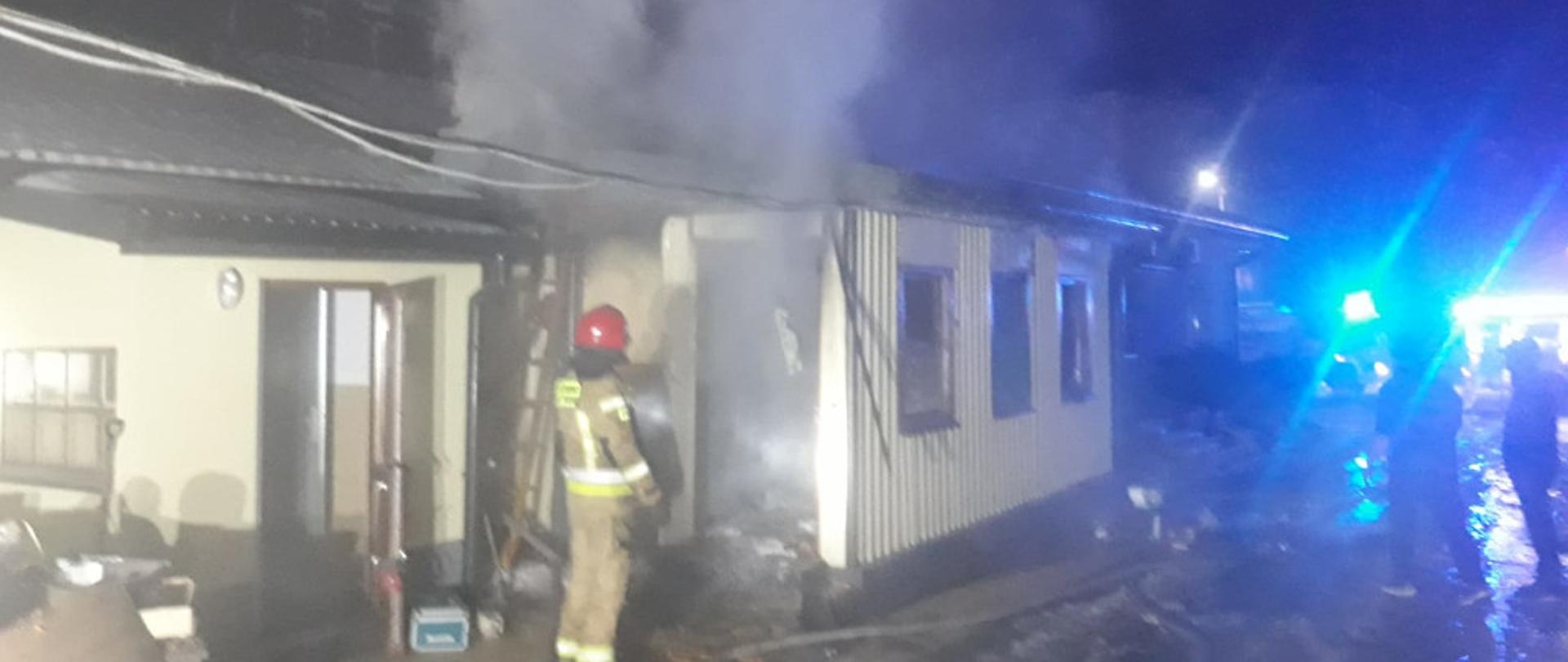 Na zdjęciu przedstawiony budynek parterowy na terenie jednej z jędrzejowskich firm. Z budynku wydobywa się intensywne zadymienie. Przed drzwiami do budynku stoi strażaków odwrócony plecami do fotografa w ubraniu specjalnym z założonym czerwonym hełmem na głowę. 