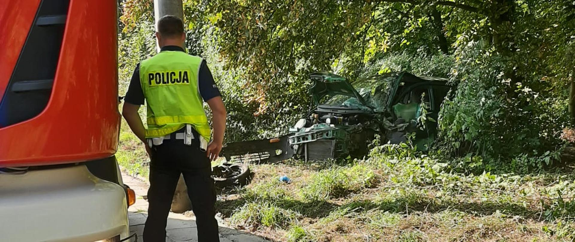 część przednia pojazdu straży pożarnej z lewej strony, w centralnej części znajduje się funkcjonariusz policji, który jest tyłem do obiektywu oraz słup żelbetonowy od lampy ulicznej. Z prawej strony samochód osobowy marki Daewoo Matiz koloru zielonego rozbity od przedniego uderzenia pojazdu w słup. Urwane koło oraz oderwany zderzak leżą na poboczu.