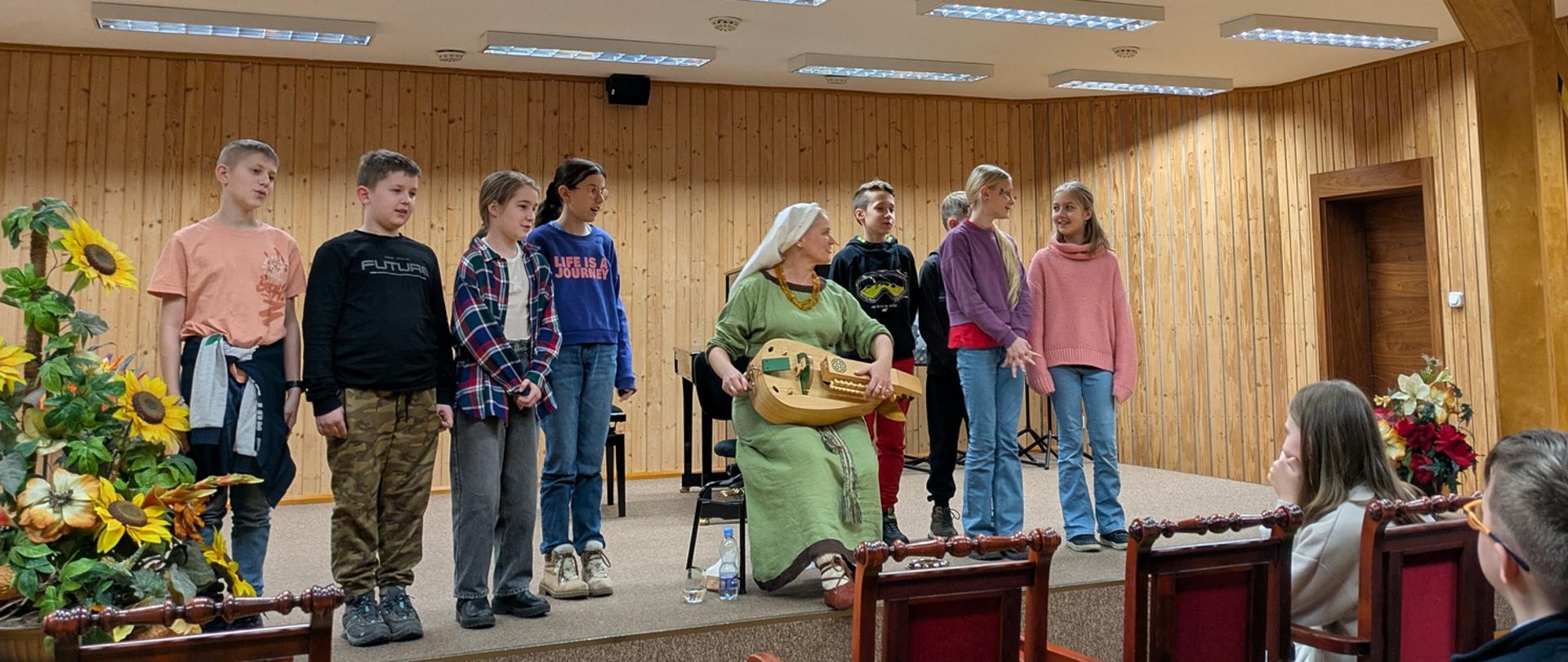 Kobieta w ludowy stroju gra na lirze i śpiewa, za nią stoi ośmioro dzieci i również śpiewają.