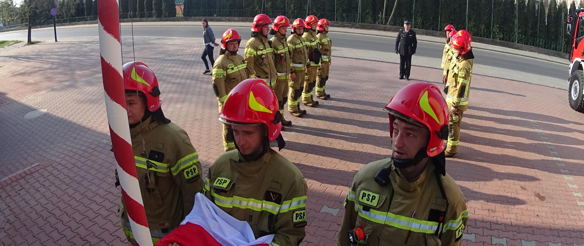 Strażacy w ubraniach specjalnych w trakcie uroczystego podnoszenia flagi na maszt.