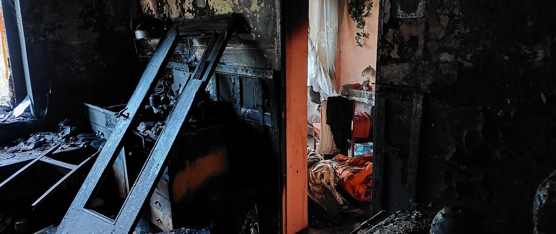 Widok na spalony pokój. Spalone całe wnetrze, ściany czarne od okopceń oraz łuszczącej się farby. W rogu pomieszczenia z lewej strony nadpalone szafki, oparte o szafki drzwi. wewnętrzne z miejsce na przeszklenie w środkowej części. Drzwi czarne, spalone. W środkowej części ściany otwór drzwiowy. W tle widać pomieszczenie, bez widocznych oznak pożaru, widać stół, krzesła, części garderoby.
